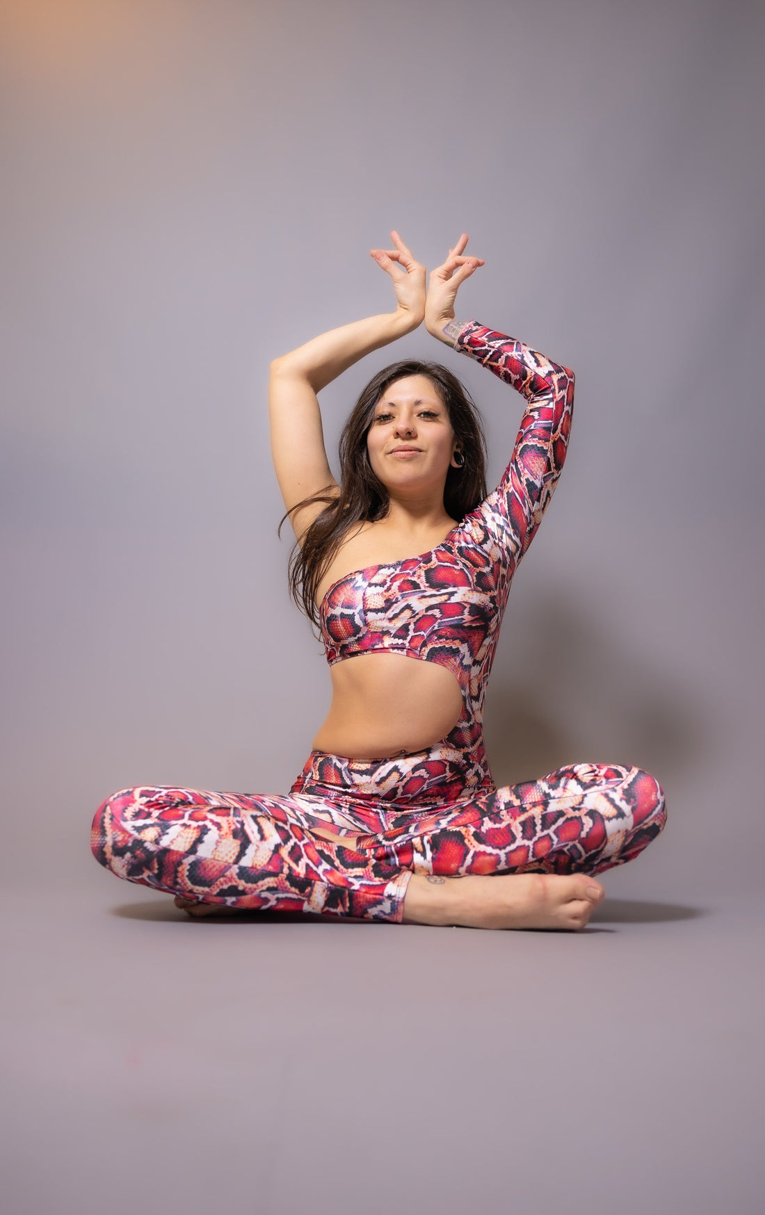 In a Red Snake-Print Isolated Catsuit with a waist cutout, crafted from Lycra, an individual elegantly executes a seated yoga pose against a plain backdrop, highlighting versatile performance wear.