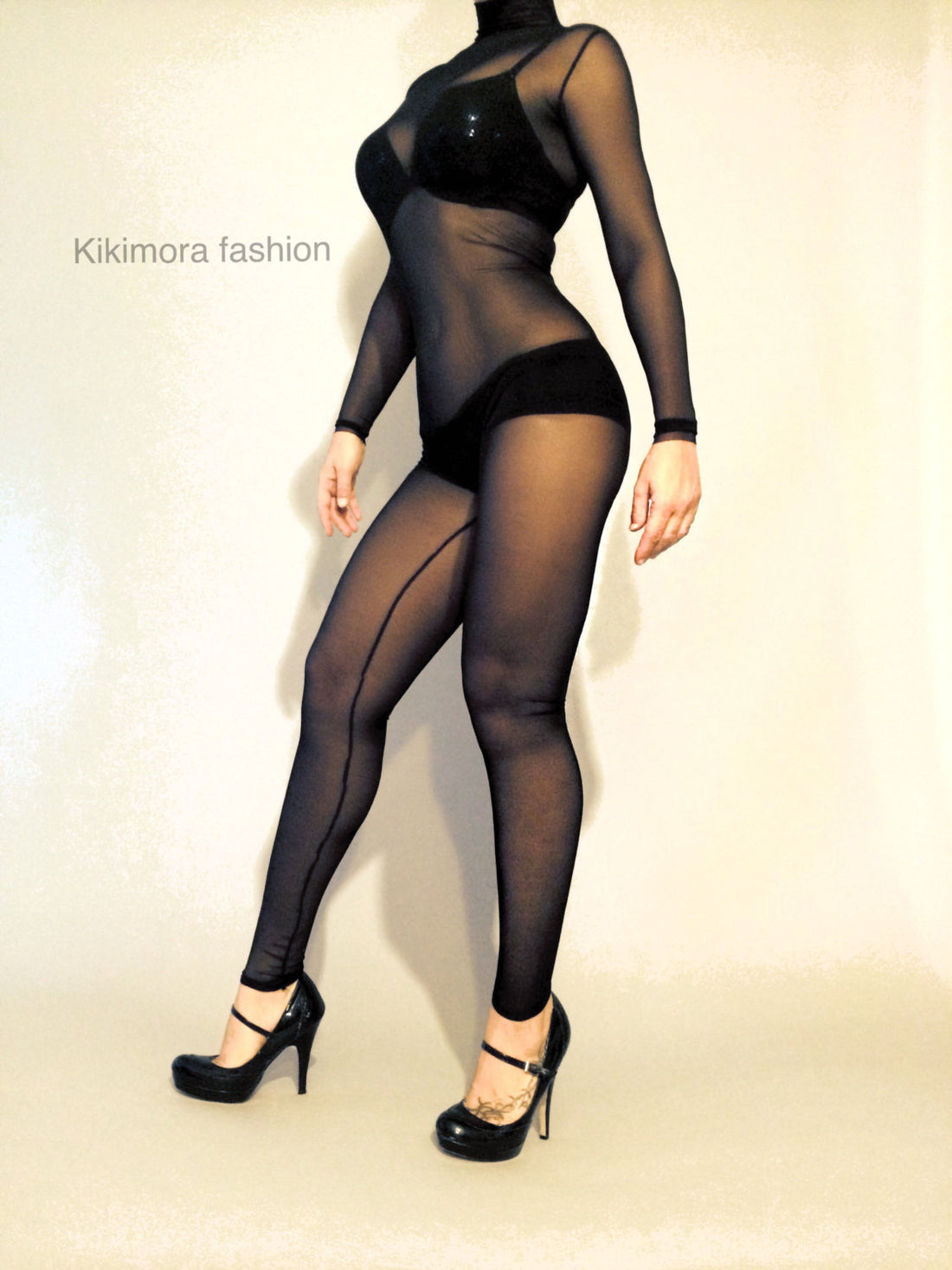 Striking a pose against a plain backdrop, a person showcases the versatility of the Black Sheer Catsuit Bodysuit with full sleeves and turtleneck, paired elegantly with heels.